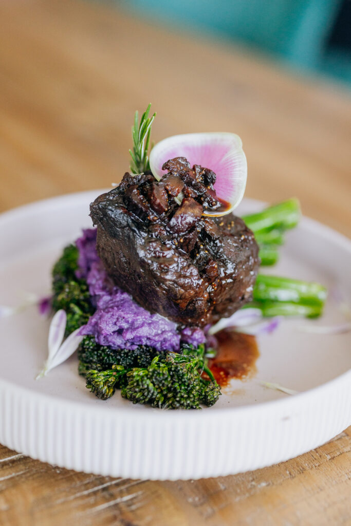 Short rib on top of purple and broccolini on a pink plate on a wooden table for event catering in Los Angeles and event catering in Orange County