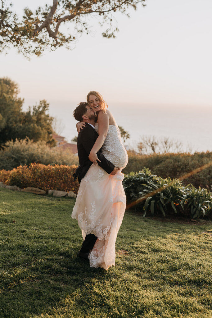 Golden Hour Wedding Photo with Pacific Ocean and Grass Lawn and Hedges with Groom Holding Up Bride