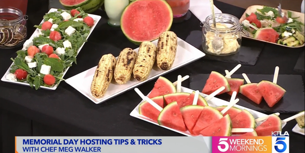 Screenshot of KTLA Memorial Day segment featuring Chef Meg Walker of Made by Meg Catering, including the grilled corn on the cob with house made honey butter and Maldon salt in the center of the photo, in between a watermelon salad and watermelon popsicles. Four pieces of corn are on a white plate on a black tablecloth. The watermelon salad on the left has watermelon balls, feta cheese, mint leaves, & arugula with lemon vinaigrette.
