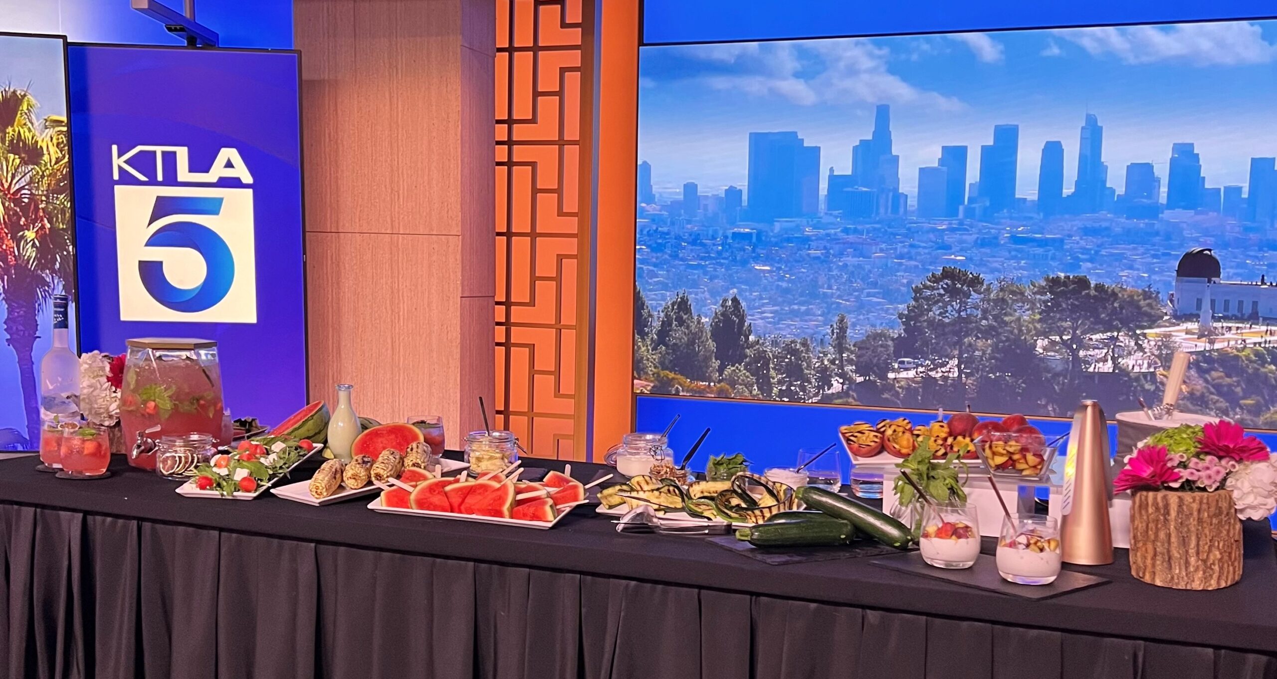 Table at KTLA 5 studio with summer items on a black tablecloth, including florals, watermelon mocktail, watermelon salad, corn on the cob, watermelon popsicles, grilled zucchini and ricotta, and grilled peaches with vanilla ice cream and balsamic reduction.