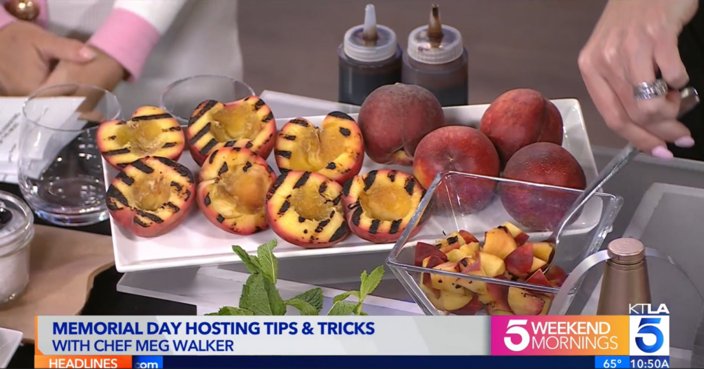 Screenshot of KTLA Memorial Day segment featuring Chef Meg Walker of Made by Meg Catering with grilled yellow peaches on a white plate next to whole yellow peaches, in front of two squeeze bottles filled with balsamic reduction. Hand going into glass bowl filled with pre-cut grilled yellow peaches. 