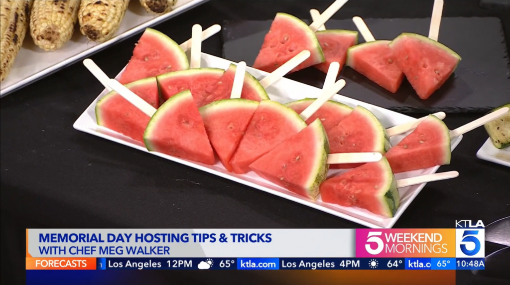 Screenshot of KTLA Memorial Day segment featuring Chef Meg Walker of Made by Meg Catering with chilled watermelon popsicles, watermelons cut in triangles with a popsicle stick in the rind on a plate on top of a black tablecloth.