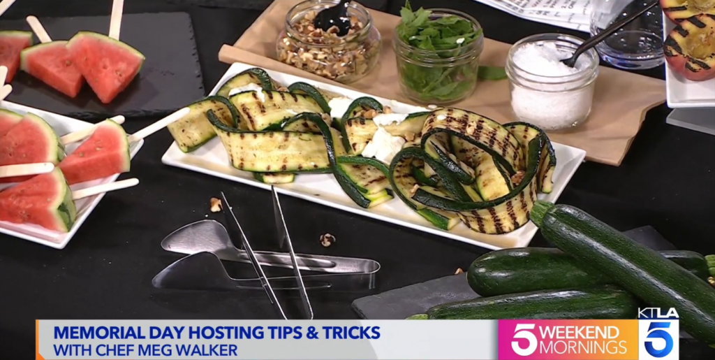 Alt Text: Screenshot of KTLA Memorial Day segment featuring Chef Meg Walker of Made by Meg Catering, including the grilled zucchini with ricotta cheese in the center of the table on a white plate. Behind it are three glass jars filled with walnuts, fresh mint, and Maldon salt. 
