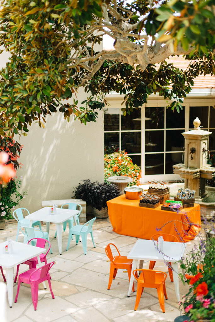 Colorful backyard table setup for children's party with light blue chairs, orange chairs, pink chairs, and an orange tablecloth on the dessert table 