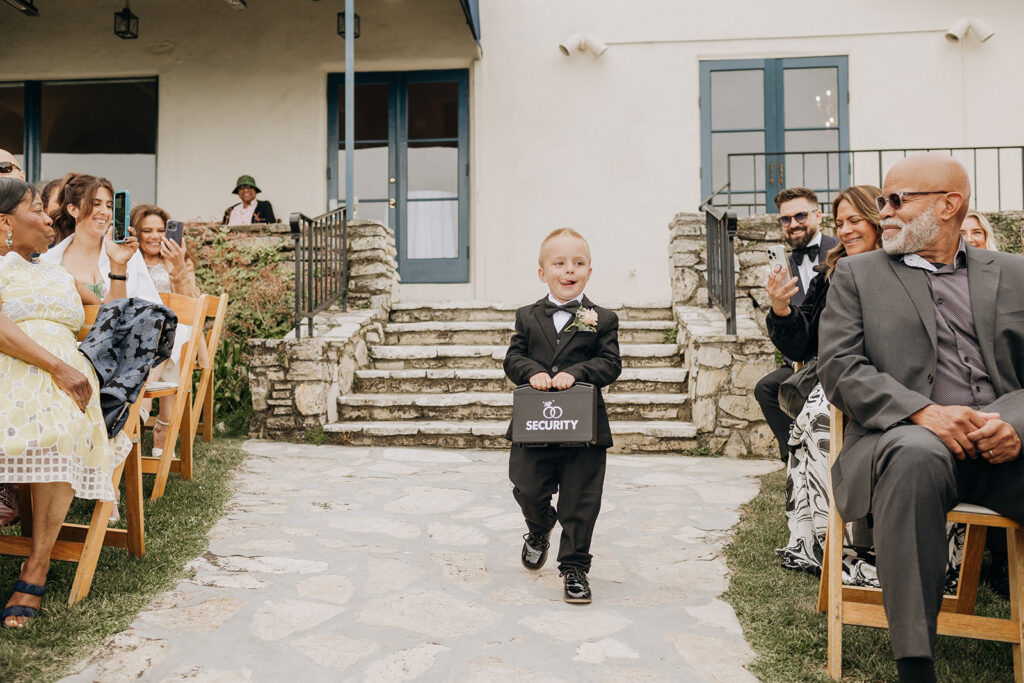 Ring Security serves as the modern Ring Bearer Walks Down Aisle Holding Black Case with Wedding Rings