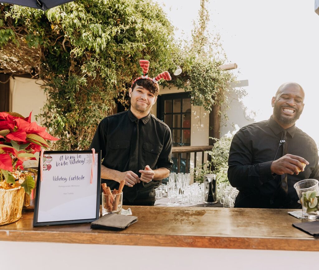 Two male bartenders in black button up shirts and black ties ready to make a Pomegranate Mimosa or Hot Toddy cocktail at the outdoor bar at A Very La Venta Holiday lunch event at La Venta Inn in Palos Verdes. 