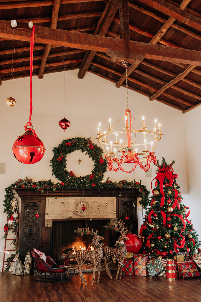 Festive Holiday Decor surrounding the fireplace in the grand ballroom at La Venta Inn in Palos Verdes. There is red beaded garland hanging from the golden chandelier, and a bell tied to the center. There are large red sleigh bells hung from the ceiling and on the floor behind the presents under the traditional Christmas tree decorated with red ribbon, lights, and red and gold ornaments. Red and gold ornaments are hung on strings of differing lengths from the fireplace mantle.  There are two wooden reindeer pulling a small red and black sleigh in front of the fireplace. 