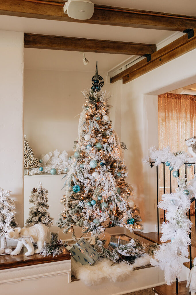 Festive Winter Wonderland Holiday Decor in the Garden Dining Room at La Venta Inn in Palos Verdes during the A Very La Venta Holiday lunch event. The frosted white and blue Christmas tree is surrounded by smaller frosted Christmas trees, a polar bear, and white garland. Off to the right in the background, you can see a rose gold photo curtain backdrop from the photobooth. 