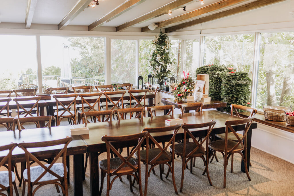 Long wooden tables with many wooden chairs tucked in set up for wreath making workshop at A Very La Venta Holiday party as a family activity at La Venta Inn in Palos Verdes. Garden dining rooms are converted into craft activity rooms with lots of natural light from the large windows facing the garden. 