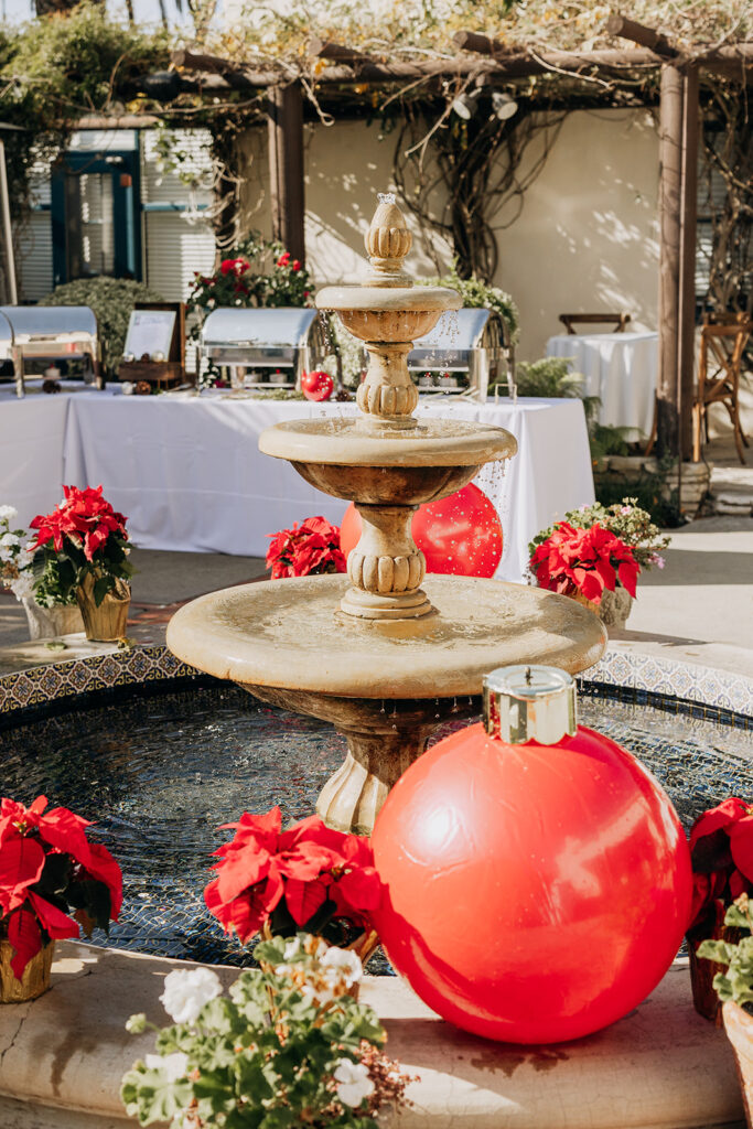 Historic Fountain Decorated at La Venta Inn in Palos Verdes with giant red ornaments and multiple red poinsettias at the base of the fountain, all in natural sunlight outside. Behind the fountain, you can see silver chafing serving dishes with warmers on a table with white tablecloths 