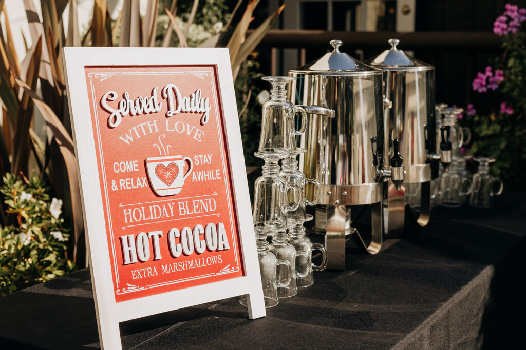 Sign for Outdoor Hot Chocolate Beverage Station with Extra Marshmallows at La Venta Inn in Palos Verdes with two metal caraffes on a black linen tablecloth next to clear hot mugs, all in natural sunlight outside