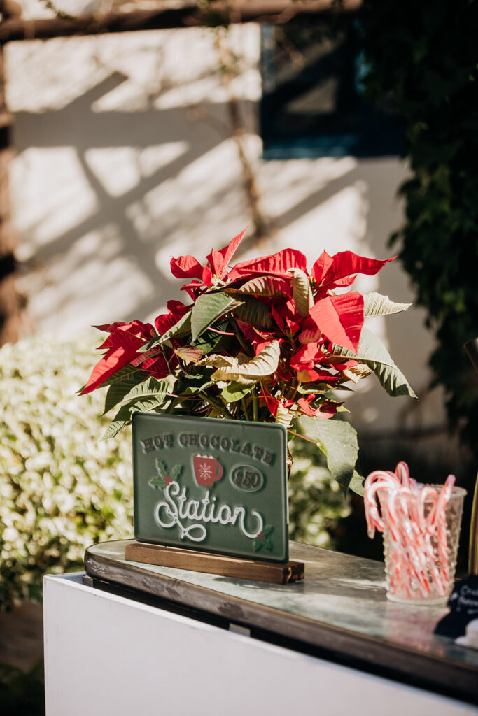 Sign for Outdoor Hot Chocolate Beverage Station at La Venta Inn in Palos Verdes with a red poinsettia on the bar and a glass with candy canes, all in natural sunlight outside