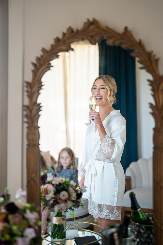 Reflection of the bride raising a glass in the mirror wearing a white bath robe.