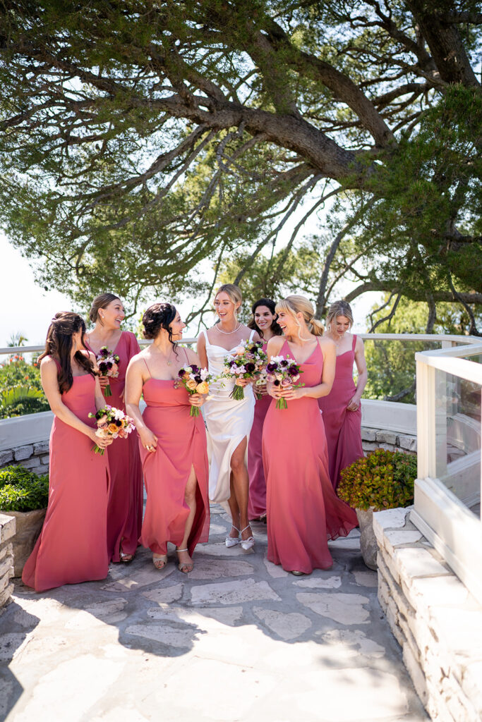 The bride takes a picture with her bridesmaids wearing pink dresses and holding floral bouquets. 