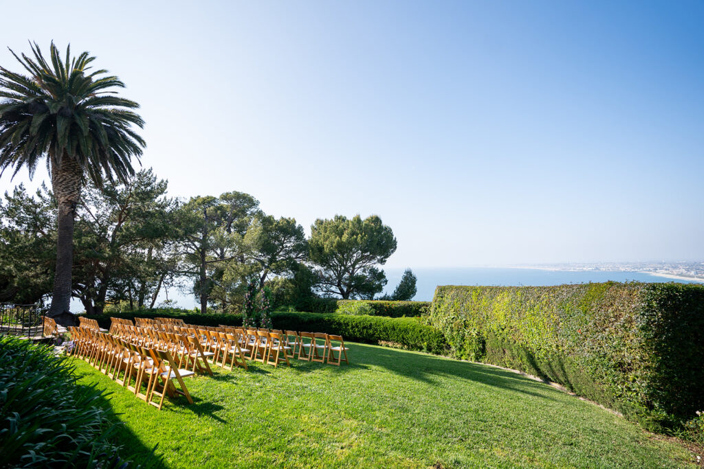 Outdoor ocean view wedding set-up with blue skies, green hedges and brown chairs in the background.