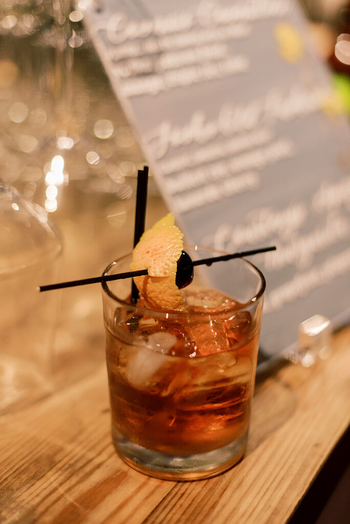A close up shot of an old fashion drink consisting of: simple syrup, luxury cherries, orange peel and bitters with two tiny black straws.