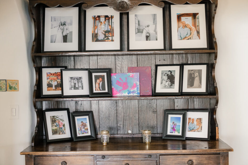 A wide angle shot of picture frames filled with family pictures on a wood shelf. In the middle is a canvas of a pink hand fingerprint.