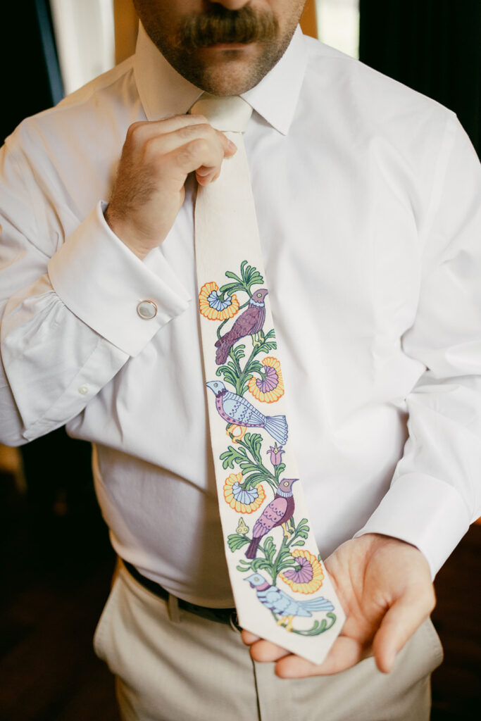 A guy is holding up a custom beige tie with yellow and green florals that is accompanied by blue and purple birds. He is wearing a white button up and cream pants.