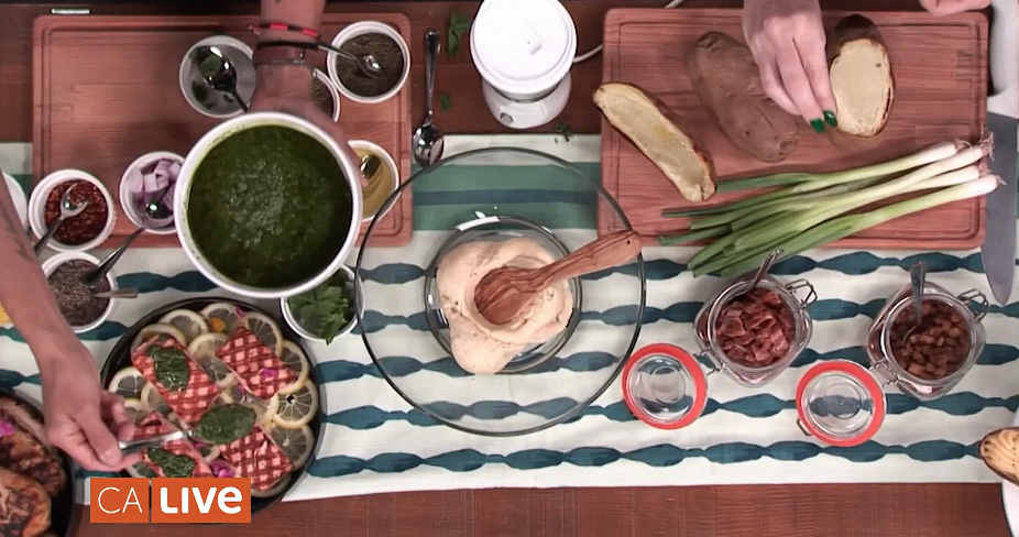 Close up shot of the table with the dishes. The table consists of a brown background and a blue and white table cover. The food varies from: salmon with lemon and chimichurri sauce, potatoes, asparagus and side ingredients.