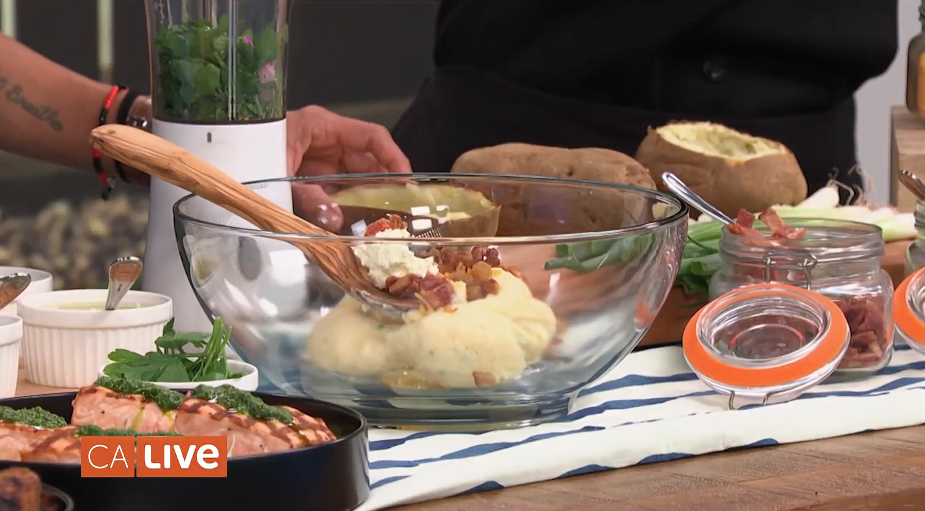 A close-up view of mashed potatoes in a clear bowl about to be mixed with butter and bacon.