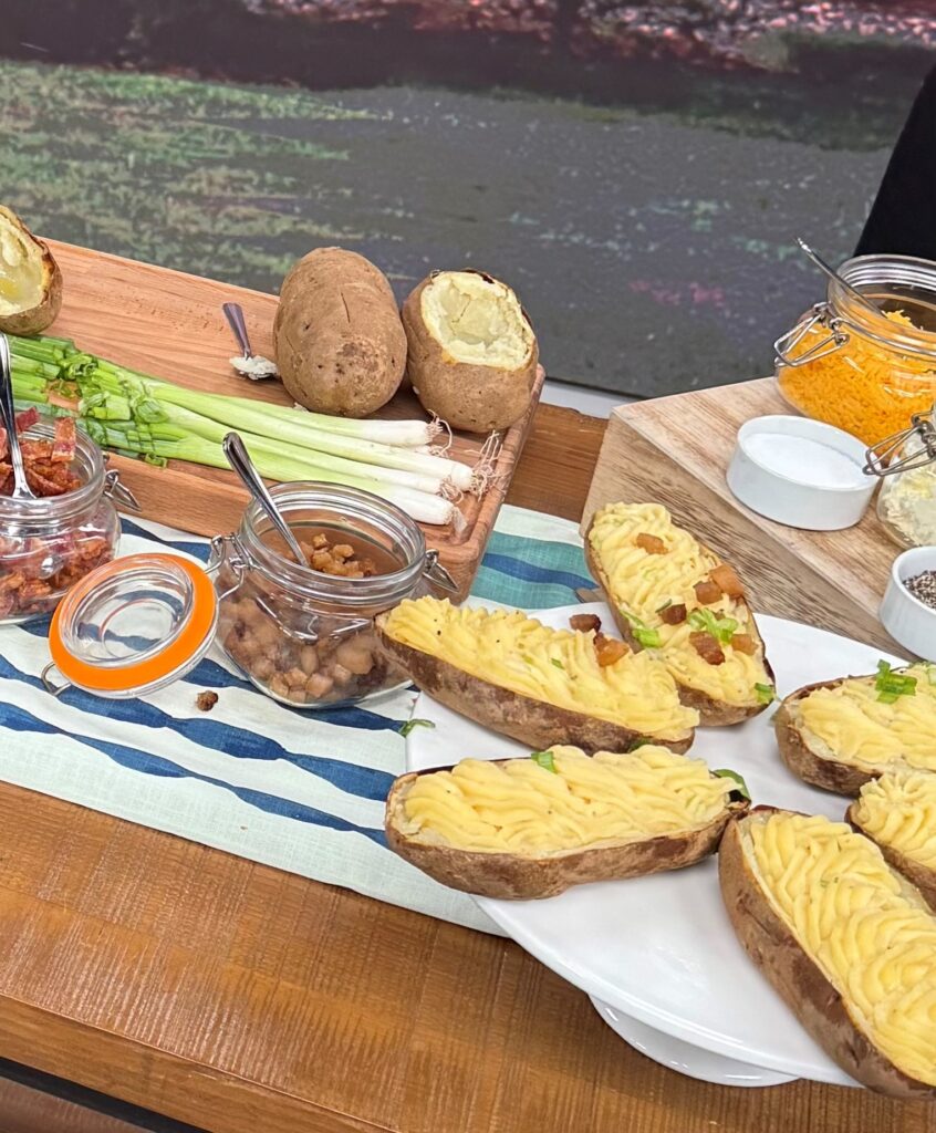 Another close up shot of the mashed potatoes on the table. On the right corner, there are green onions and potatoes on a cooking board. Next to the mashed potatoes are bacon garnishes.