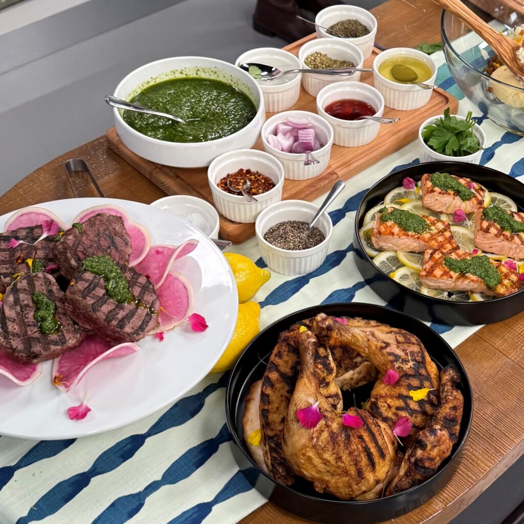 Another view of the table consisting of steak, chicken and steak garnished with chimichurri. The table is also surrounded by side ingredients such as onions, red pepper flakes, salt and pepper, oil, etc.