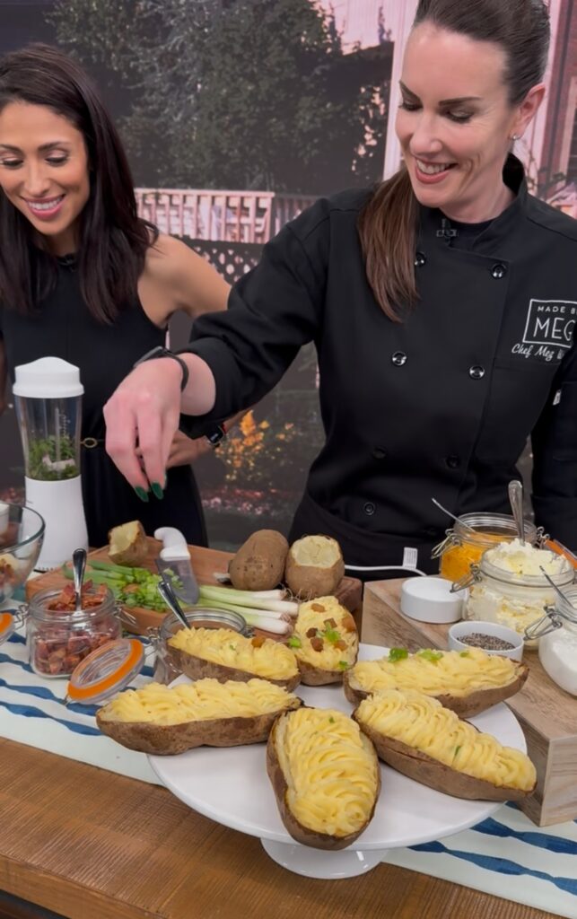 An action shot of Meg sprinkling green onions on top of mashed potatoes inside the potato skin. The surrounding contains several potatoes, bacon, sour cream, butter and a blender.