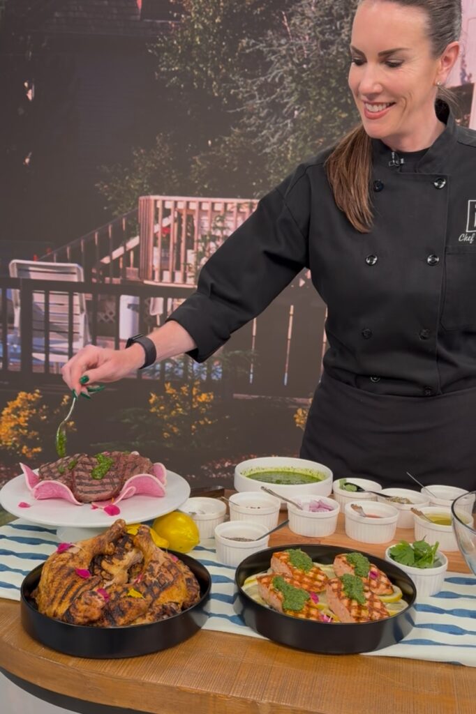 An action shot of Chef Meg garnishing the steak with chimichurri. The table also consists of chicken and steak garnished with chimichurri. 