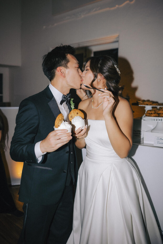 An action shot of the bride and groom kissing each other while holding a cup of ice cream with a chocolate chip cookie on top.