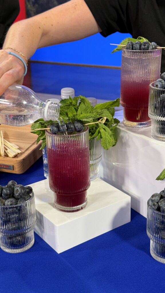 A closer shot of the action view of the lady pouring the club soda on top of the mocktail drink. The cup contains a blueberry flavored drink, topped with fresh mint leaves and blueberries on a skewer stick.