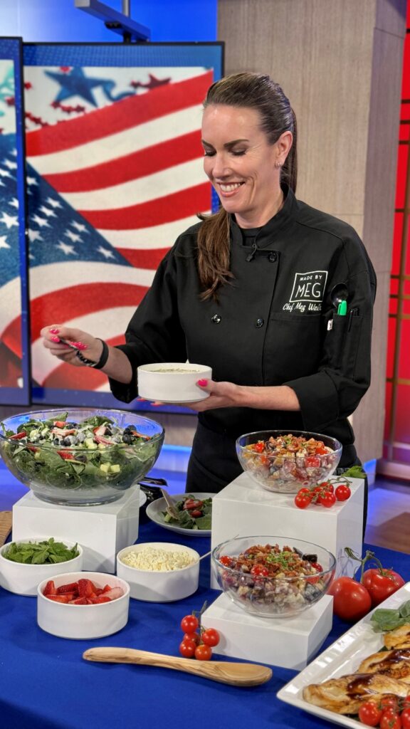 Action shot of Chef Meg, holding a bowl and putting toppings on the salad. The table consists of three main salad bowls on white risers surrounded by smaller bowls of blue cheese, strawberries, tomatoes and kale. Behind Chef Meg is an image of an American flag.