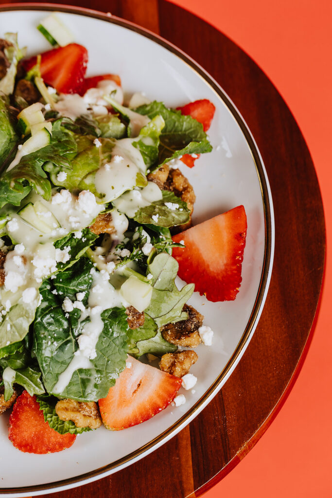 Close - up shot of a baby kale salad with strawberries and dressing in a white plate with a red background.