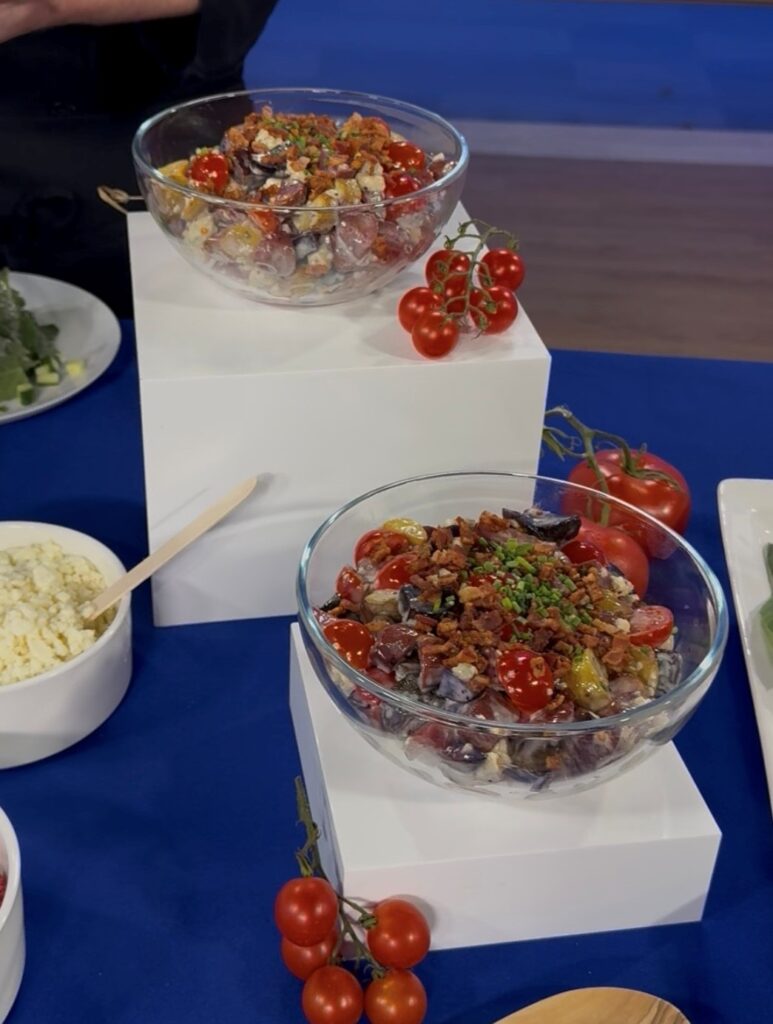 Another close-up view of two main salad bowls both on white risers surrounded by cherry tomatoes on vines. Both salad contains tri-colored baby potatoes with potato skins left on, cherry tomatoes, crumbled blue cheese, crisp bacon, thinly sliced chives and a drizzle of blue cheese dressing.