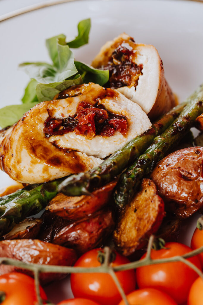 Close up shot of a savory stuffed chicken breast on a white plate. The stuffings contains: creamy goat cheese, sun-dried tomatoes, fresh basil and marsala wine sauce topping. It is accompanied by fresh basil, honey potatoes and tomatoes on the vine.
