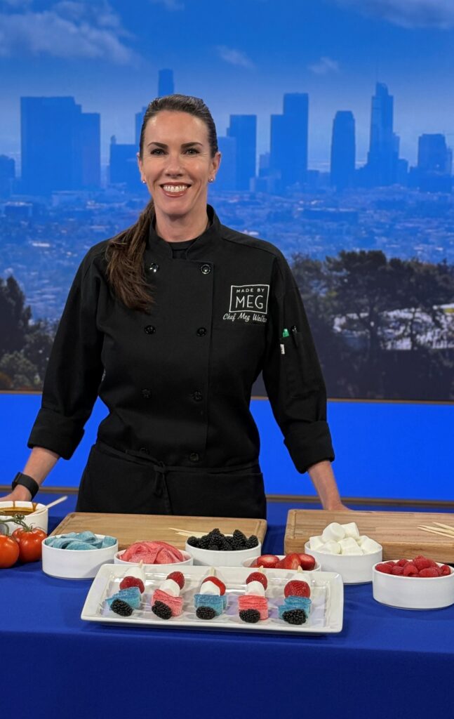 A candid photo of Chef Meg with a table filled with desserts. The table consists of several white bowls containing: red and blue flat sour belts, blueberries, strawberries, raspberries and marshmallow. In front of the bowls is a plate filled with fruit and candy kebabs with the ingredients mentioned previously. 