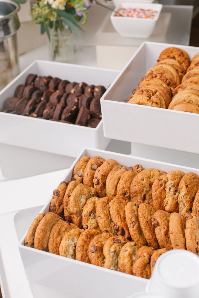 A candid shot of the cookies, all in white trays. On the left corner is white chocolate chip macadamia nut cookies and oatmeal raisin cookies on the right. On the bottom right corner are chocolate chip cookies.