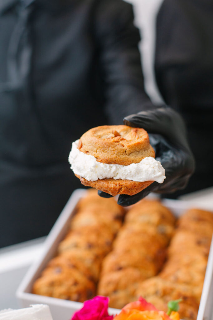 Close up shot an ice cream sandwich being held by a server who is wearing black gloves. In the background is a white tray filled with cookies.