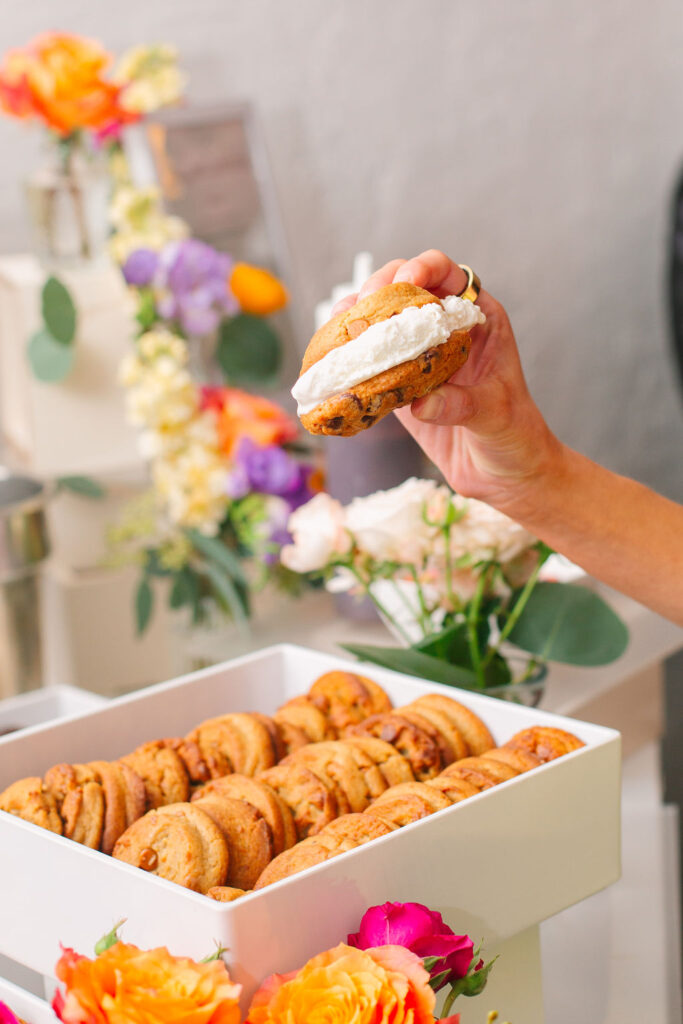 Close up shot of someone holding an ice cream sandwich. In the background, there is a white tray filled with cookies and a floral arrangement as decorations.