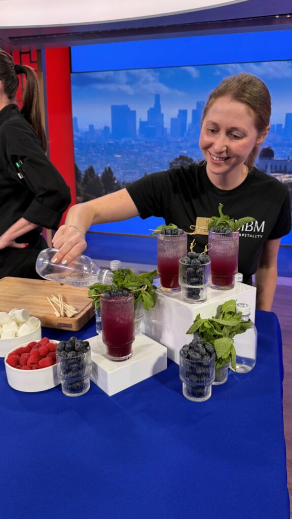 An action shot of a lady pouring club soda on top of the blueberry lemon fizz mocktail. The mocktail is topped with fresh mint leaves and blueberries on a skewer sticks. The image contains several cups of the drink, others are filled with blueberries. The table is also surrounded by fresh mint leaves and blueberries.