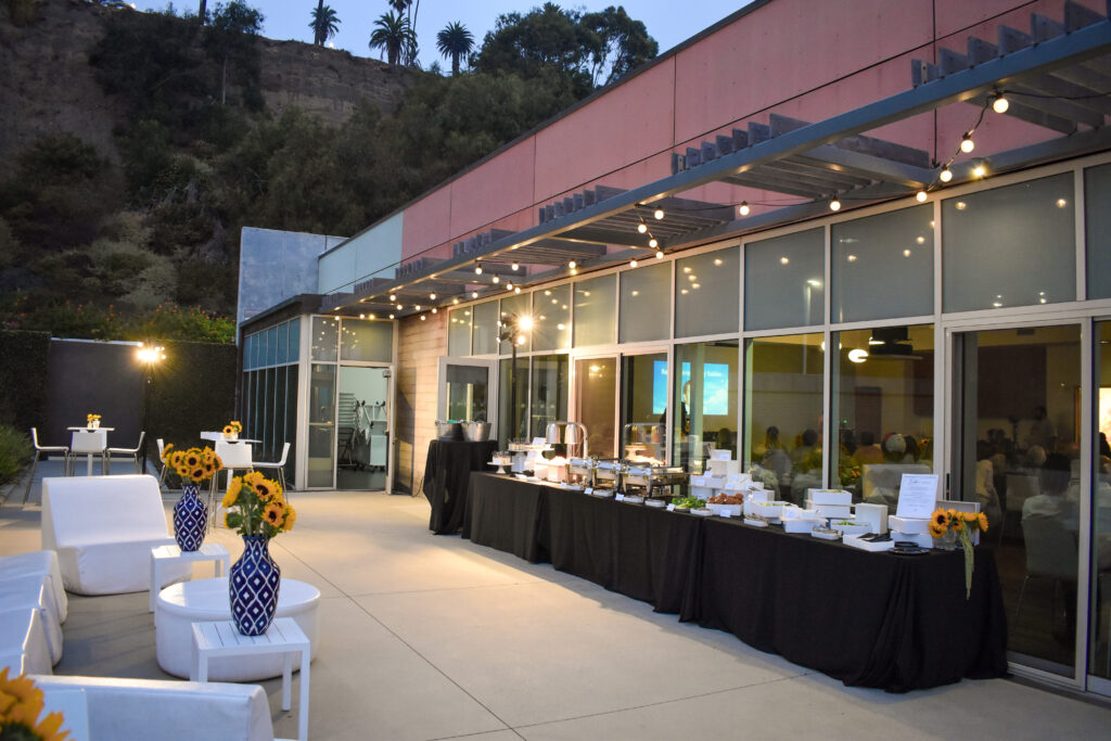 A stunning evening setup at the Annenberg Beach House, featuring an elegant outdoor buffet under a modern building with pink and blue walls, illuminated by string lights and spotlights. The long black-draped table showcases chafing dishes with warm dishes, white trays of appetizers, and floral centerpieces with yellow sunflowers in blue patterned vases, creating a sophisticated ambiance for events. White modern lounge furniture with round tables is scattered in the foreground, offering a cozy seating area with views of the lush hillside and palm trees in the background. Large glass doors reveal an indoor space with guests and a screen, ideal for corporate events or weddings, enhancing the venue’s appeal.