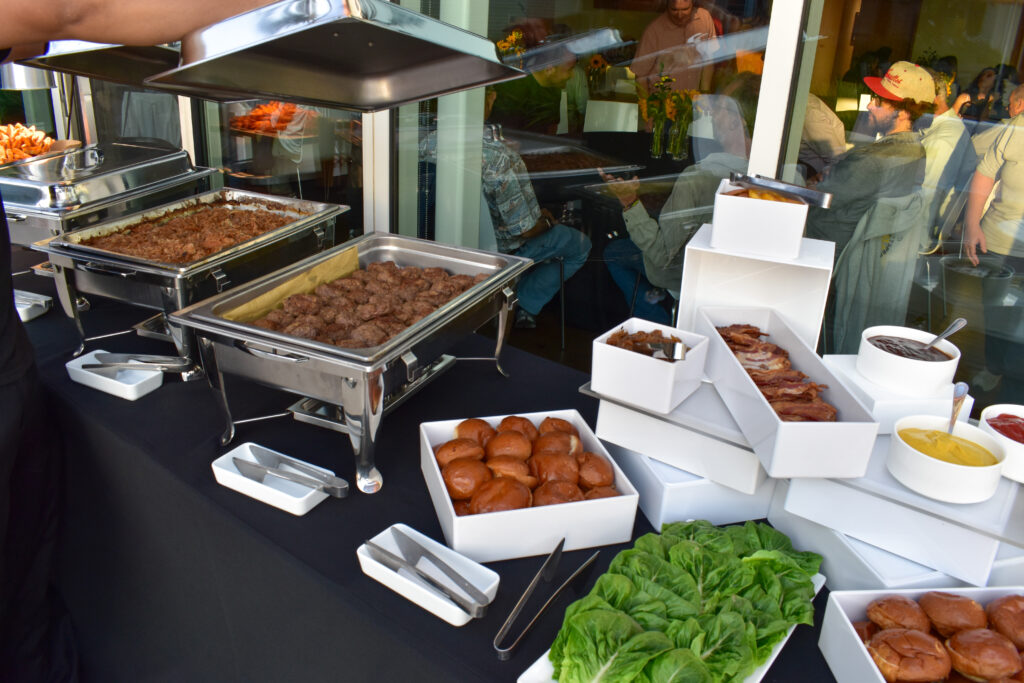 A gourmet burger bar catering station by Made by Meg Catering at the Annenberg Beach House, featuring an elegant black tablecloth setup with stainless steel chafing dishes and modern white serving trays. The buffet includes trays of freshly grilled beef patties, shredded pulled beef, and golden potato wedges kept warm under heat lamps. In the foreground, a display of shiny brioche slider buns, crisp romaine lettuce leaves, and neatly arranged toppings such as crispy bacon strips and caramelized onions are presented in white square and rectangular dishes stacked on tiered stands. To the right, three white ceramic bowls hold classic burger condiments — ketchup, mustard, and barbecue sauce — each with a serving spoon. Silver tongs rest in sleek white trays for easy self-service. Behind the buffet, guests can be seen enjoying the event indoors, reflected in the glass, with warm lighting and flower arrangements creating a lively yet refined coastal atmosphere. The setup showcases Made by Meg Catering’s upscale, organized presentation perfect for weddings, corporate events, and private gatherings at the Annenberg Beach House.