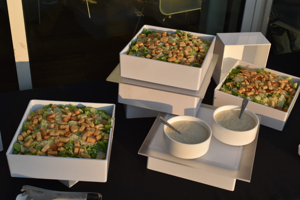 A beautifully presented buffet salad station at the Annenberg Beach House, catered by Made by Meg Catering. The display features several large, modern white square bowls filled with fresh Caesar salads topped generously with crunchy golden-brown croutons and shaved Parmesan cheese over crisp romaine lettuce. The salads are neatly elevated on tiered white risers, creating a clean, elegant, and professional catering presentation. In front of the salads sit two round white ceramic bowls filled with creamy Caesar dressing, each with a silver serving spoon. The setup is arranged on a sleek black tablecloth, with soft evening sunlight casting a warm glow across the scene. The image highlights the refined yet coastal atmosphere of an outdoor event at the Annenberg Beach House, showcasing Made by Meg Catering’s sophisticated and stylish presentation for weddings, receptions, and private gatherings.