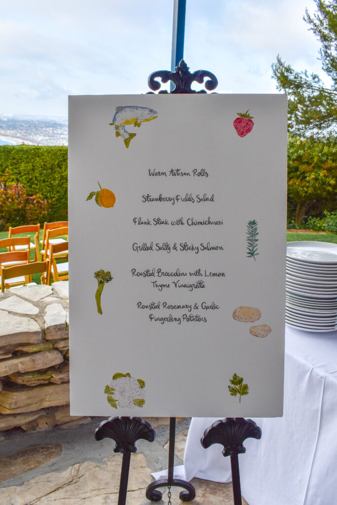 Elegant wedding buffet menu display at La Venta Inn, an ocean-view event venue in Palos Verdes Estates, California. The menu is printed on a white poster board adorned with hand-painted watercolor illustrations of ingredients, including a fish, strawberry, lemon, herbs, broccoli, potatoes, and cauliflower. The board is held upright on a black wrought-iron easel with ornate scroll details. The custom menu, catered by Made by Meg Catering, features a gourmet selection: "Warm Artisan Rolls, Strawberry Fields Salad, Flank Steak with Chimichurri, Grilled Salty & Sticky Salmon, Roasted Broccolini with Lemon Thyme Vinaigrette, and Roasted Rosemary & Garlic Fingerling Potatoes." In the background, there are rows of wooden ceremony chairs with white cushions arranged on a lush green lawn overlooking the Pacific Ocean, framed by hedges and trees under a bright, partly cloudy sky. A table with stacked white plates is visible on the right side of the image, ready for guests to enjoy the meal.