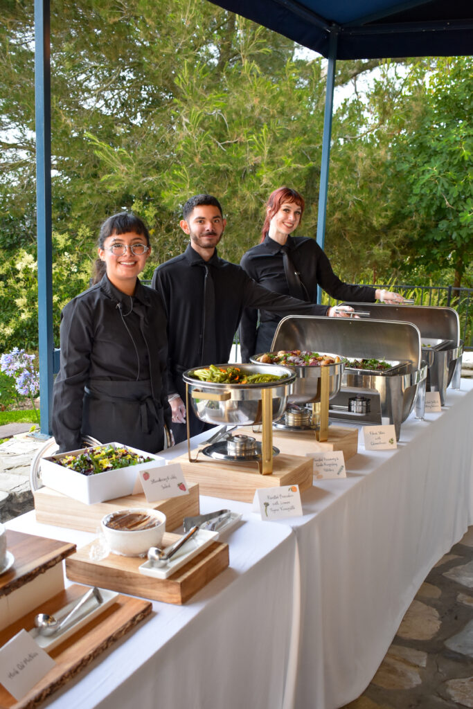 Three cheerful Made by Meg Catering staff in black uniforms serve at a lavish buffet at La Venta Inn, Palos Verdes, CA, under a blue canopy with lush greenery. The white-tablecloth setup features wooden stands with a chocolate dip bowl, strawberry salad, roasted Brussels with yuzu vinaigrette, filet mignon with chimichurri, and chafing dishes of green beans, broccoli, and roasted veggies.