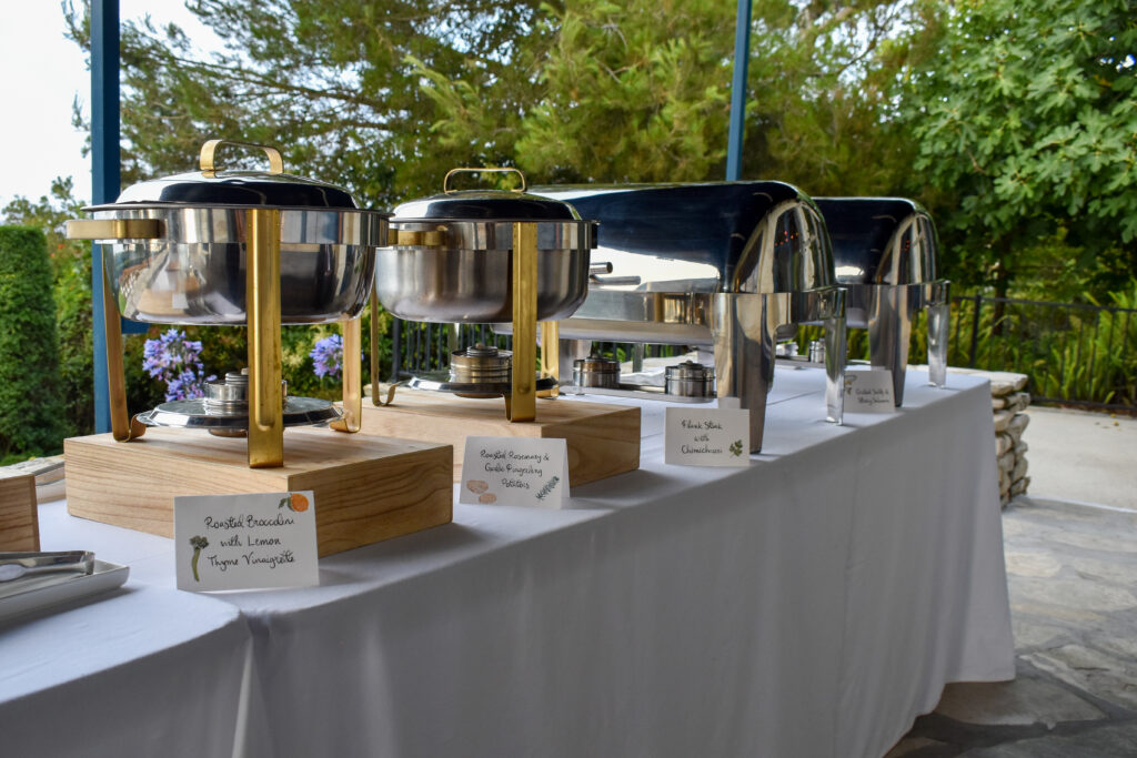 Upscale buffet station setup at La Venta Inn in Palos Verdes Estates, California, catered by Made by Meg Catering. The buffet table is covered with a crisp white tablecloth and arranged with polished stainless steel chafing dishes, some with gold accents and wooden risers for height and elegance. Each dish is labeled with custom illustrated menu cards, featuring playful watercolor-style ingredient drawings. Visible food labels include: "Roasted Broccolini with Lemon Thyme Vinaigrette," "Roasted Rosemary & Garlic Fingerling Potatoes," "Flank Steak with Chimichurri," and "Grilled Salty & Sticky Salmon." The scene is framed by lush greenery and coastal foliage under an open-air canopy, blending refined dining with a natural, ocean-view atmosphere. This elevated buffet presentation exemplifies luxury wedding and event catering by Made by Meg at the historic La Venta Inn.