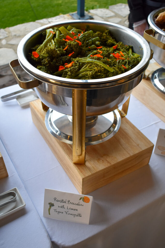 A stainless steel chafing dish at La Venta Inn showcases roasted broccolini with lemon thyme vinaigrette, catered by Made by Meg Catering, featuring vibrant green broccolini florets topped with bright orange edible flowers for a colorful presentation. The dish is elegantly displayed on a wooden base atop a white tablecloth, with a blurred outdoor backdrop of green grass and stone walls, ideal for events like weddings or corporate gatherings. A handwritten sign reading "Roasted Broccolini with Lemon Thyme Vinaigrette" with a decorative orange flower and green stem enhances the gourmet appeal.