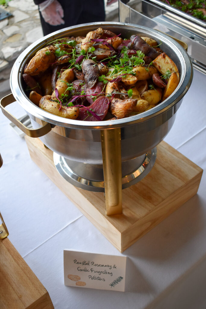 Close-up of a gourmet buffet dish served at La Venta Inn in Palos Verdes Estates, California, catered by Made by Meg Catering. The dish features a vibrant medley of Roasted Rosemary & Garlic Fingerling Potatoes, beautifully presented in a polished stainless steel chafing dish with modern gold handles, elevated on a light wood riser. The colorful fingerling potatoes—golden, red, and purple—are roasted to a crisp and garnished with fresh herbs including sprigs of rosemary, microgreens, and beet leaves, enhancing both aroma and visual appeal. A custom menu card with hand-drawn potato and herb illustrations labels the dish in elegant script. The chafing station rests on a clean white tablecloth as part of an upscale outdoor buffet spread, set against the natural stone and greenery backdrop of the ocean-view La Venta Inn wedding venue.