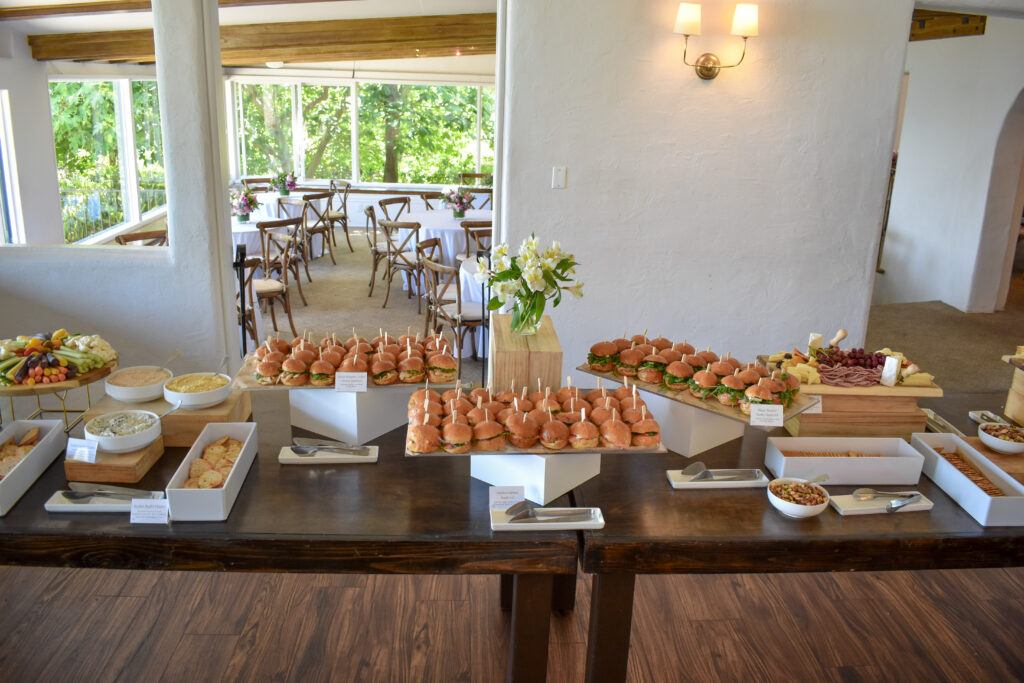 A stunning buffet setup at La Venta Inn, catered by Made by Meg Catering, for a celebration of life event, featuring a long dark wooden table laden with gourmet offerings. The spread includes mini sliders on skewers with fresh greens, a charcuterie board with assorted meats, cheeses, grapes, and crackers, and a veggie platter with colorful carrots, celery, and dips like hummus and ranch. Additional dishes include kale salad, breadsticks, and a bowl of mixed grains, all elegantly displayed with white trays and wooden stands, accented by a centerpiece of white flowers. The indoor venue boasts large windows with lush green views, rustic wooden beams, and cross-back chairs with white tablecloths in the background, creating a serene ambiance for memorial catering.