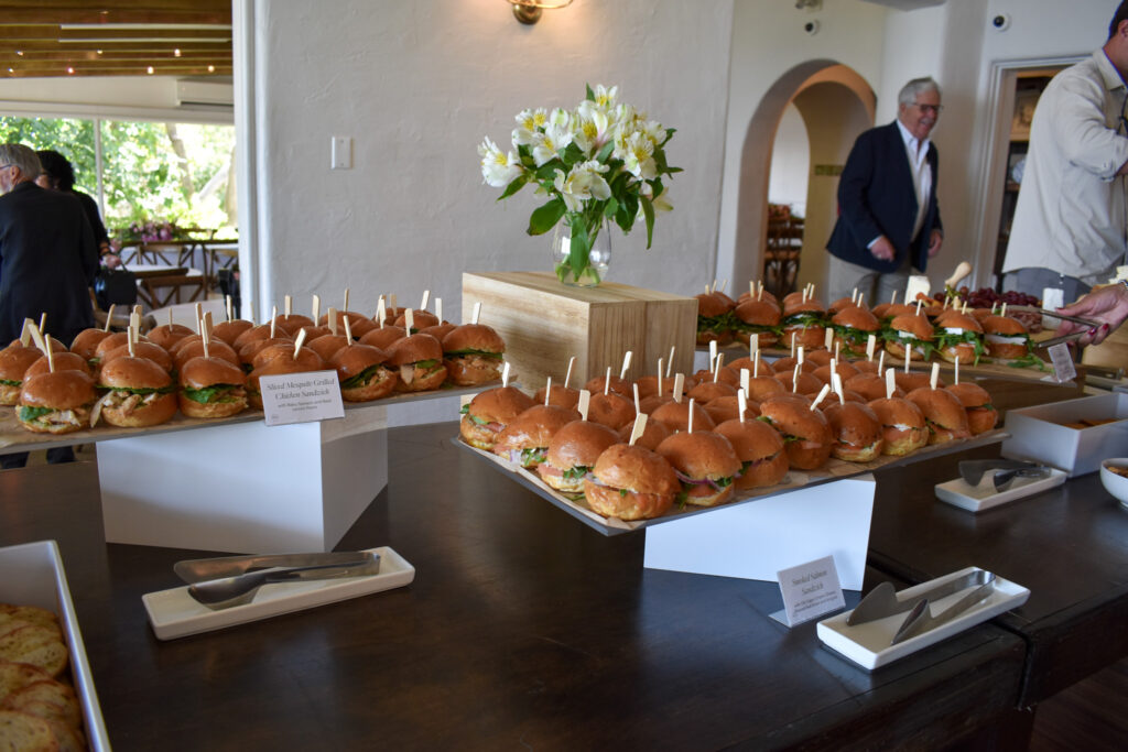 Elegant sandwich catering display by Made by Meg Catering at La Venta Inn in Palos Verdes, featuring rows of gourmet mini sandwiches beautifully arranged on white square platters. The setup includes mesquite-grilled chicken sandwiches and smoked salmon sandwiches on soft brioche slider buns, each neatly secured with a wooden skewer for easy serving. The sliders are garnished with crisp lettuce and fresh ingredients, reflecting Made by Meg’s signature California-inspired presentation. A small printed label card identifies each sandwich type, while sleek stainless steel serving tongs rest in modern white ceramic trays. The platters are elevated on minimalist white risers and a wooden crate centerpiece topped with a clear vase of white flowers, adding a touch of natural elegance. In the background, the bright and airy interior of La Venta Inn features white stucco walls, wood-beamed ceilings, and large windows overlooking lush greenery—creating a sophisticated coastal atmosphere perfect for weddings, celebrations of life, and private events. This display highlights Made by Meg Catering’s attention to detail, fresh seasonal ingredients, and upscale presentation at La Venta Inn.